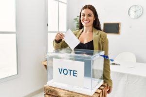 young beautiful hispanic woman smiling confident putting vote on ballot box at electoral college