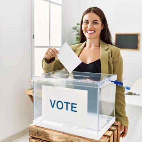 young beautiful hispanic woman smiling confident putting vote on ballot box at electoral college
